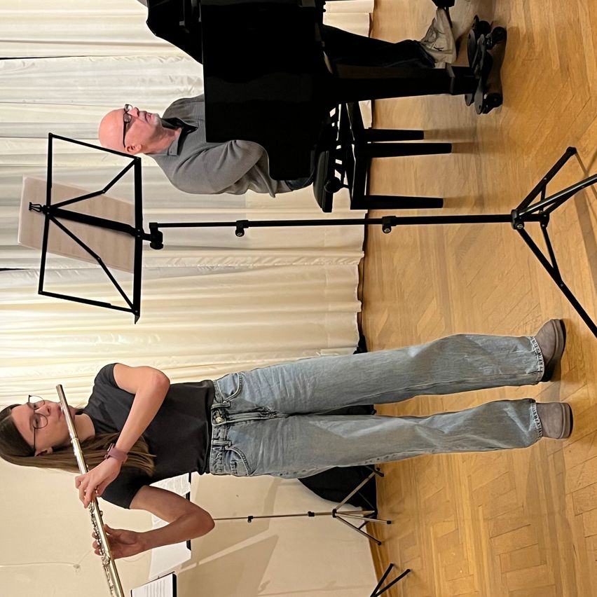 Eine Frau spielt Flöte, während ein Mann sie am Klavier begleitet. Beide stehen in einem Raum mit Holzboden und weißen Vorhängen im Hintergrund.