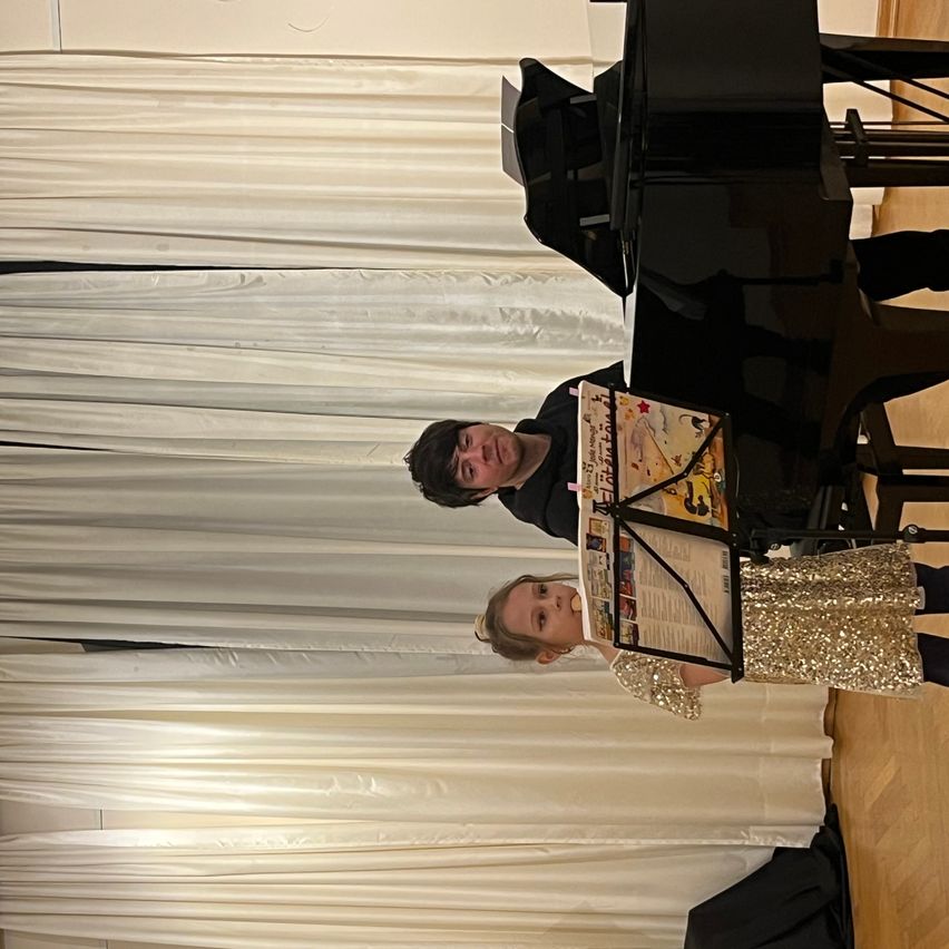 Ein Mann und ein junges Mädchen stehen vor einem schwarzen Klavier. Das Mädchen hält ein Notenbuch, und der Mann schaut sie an. Hinter ihnen hängen weiße Vorhänge von der Decke.