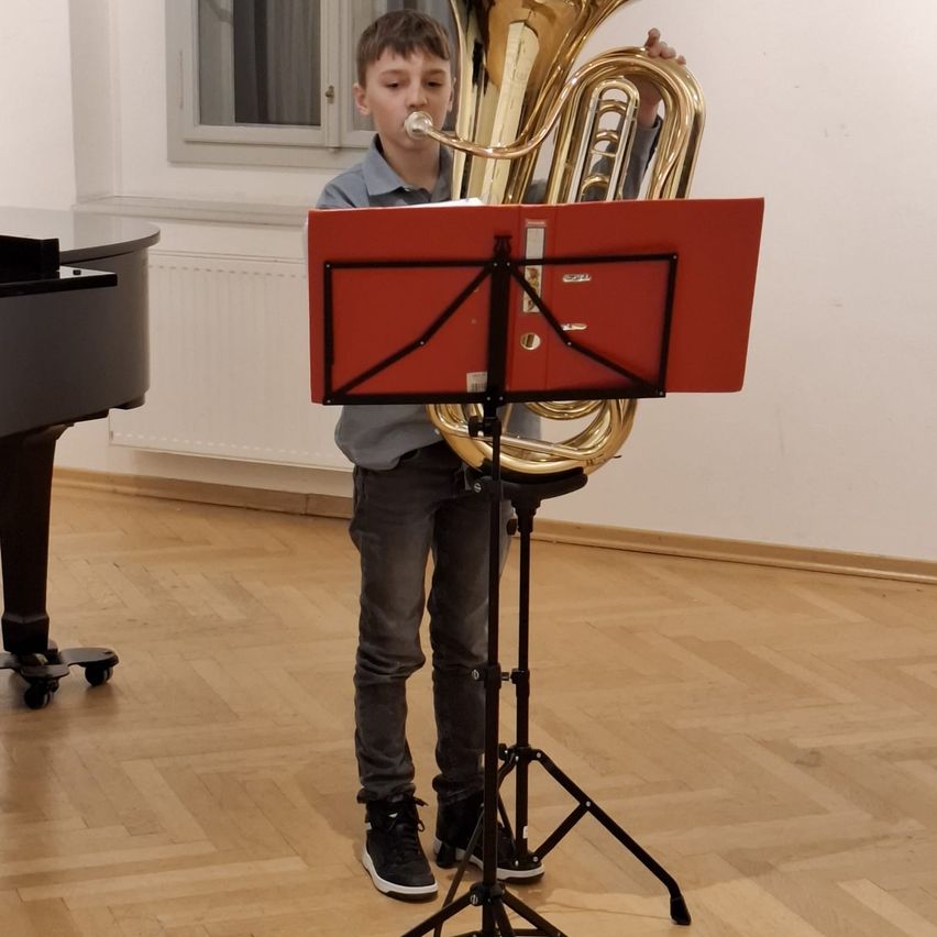 Ein Junge spielt eine Tuba, während er in einem Raum steht. Ein Notenständer mit Noten steht vor ihm, und ein Klavier befindet sich hinter ihm.