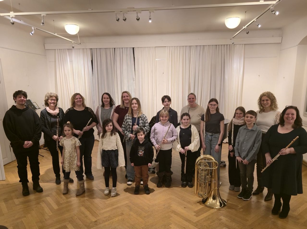 Eine Gruppe von Menschen, wahrscheinlich eine Musikband, steht auf einem Holzboden. Sie halten verschiedene Musikinstrumente. Einige halten Flöten, während andere Trompeten und Tuben haben. Der Raum hat weiße Vorhänge und eine goldene Tuba in der Mitte.