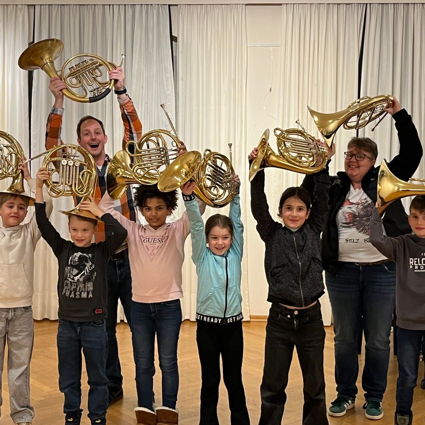 Eine Gruppe von Kindern und zwei Erwachsenen halten französische Hörner hoch. Sie lächeln und posieren für ein Foto in einem Raum mit weißen Vorhängen.