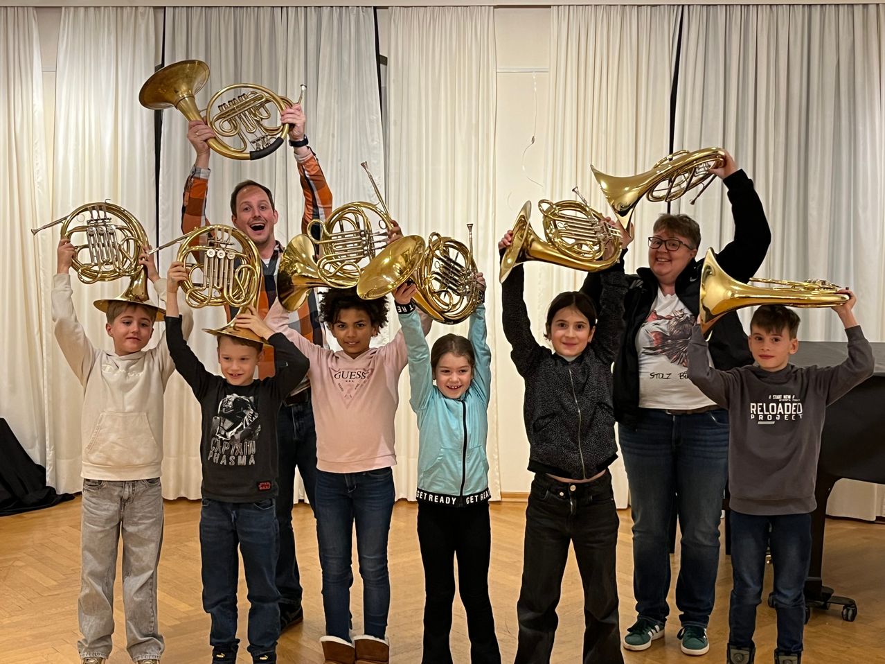 Eine Gruppe von Kindern und zwei Erwachsenen halten französische Hörner hoch. Sie lächeln und posieren für ein Foto in einem Raum mit weißen Vorhängen.