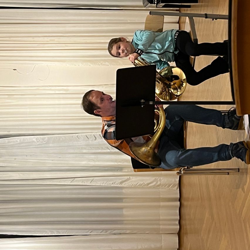 Ein Musiker spielt ein Blechblasinstrument, während ein Kind zusieht. Sie sitzen auf Stühlen in einem Raum mit Holzböden und weißen Vorhängen.