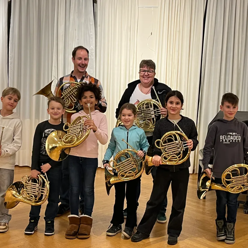 Eine Gruppe von Kindern und zwei Erwachsenen lächelt und posiert für ein Foto, während sie französische Hörner halten. Sie befinden sich in einem Raum mit weißen Vorhängen und Holzböden.