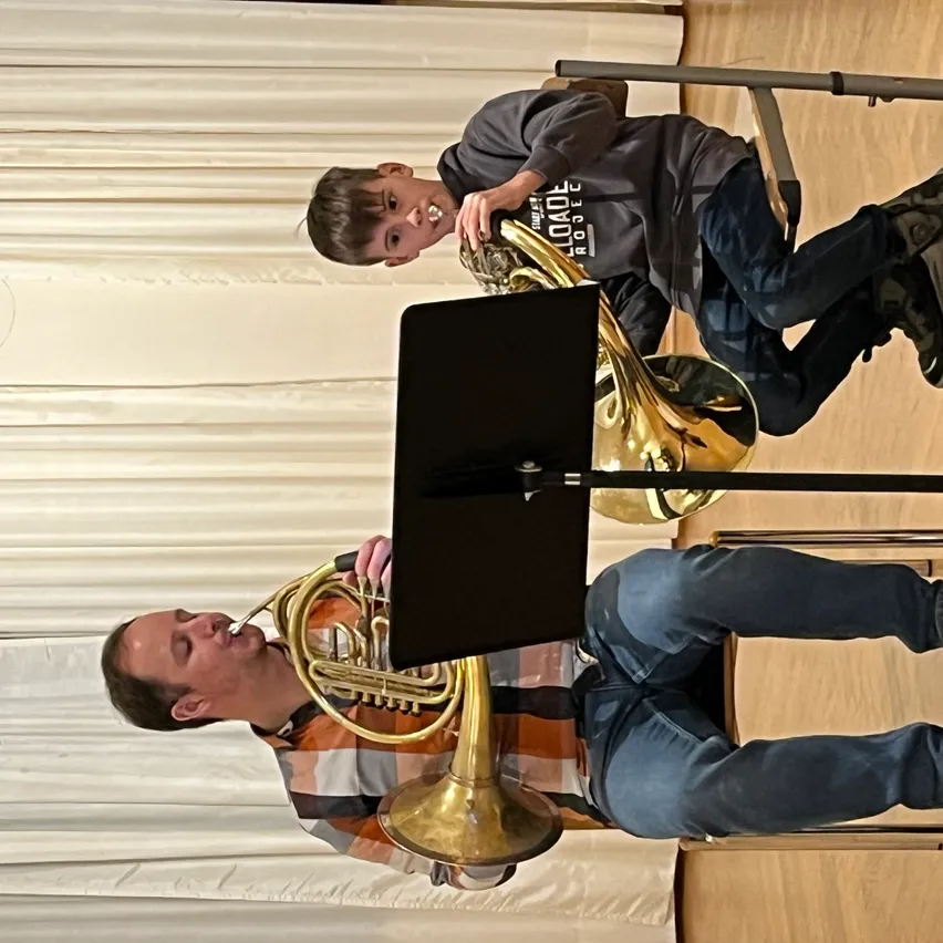 Zwei Personen spielen ein Blechblasinstrument. Einer ist ein junger Junge, der hinter einem sitzenden Mann steht, beide konzentrieren sich auf ihre Instrumente.