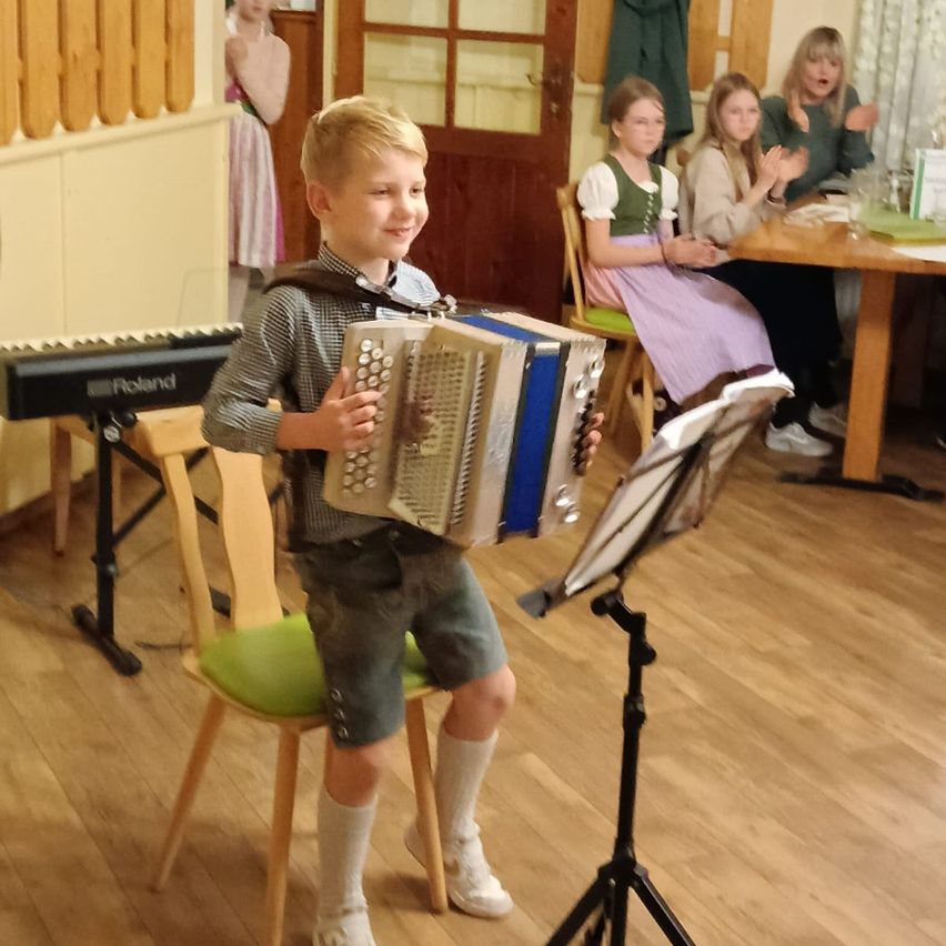 Ein junger Junge spielt Akkordeon sitzend auf einem Stuhl, mit einem Tisch, Stühlen und einem Klavier im Hintergrund. Einige Leute sitzen und klatschen.
