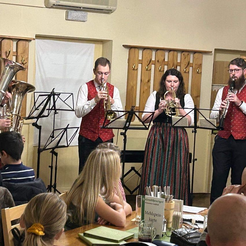Eine Band spielt Trompeten in einem Raum mit Holzwänden und einem Tisch. Ein Schild auf dem Tisch steht Mehlspeise Karte. Zuhörer sitzen herum und lauschen.