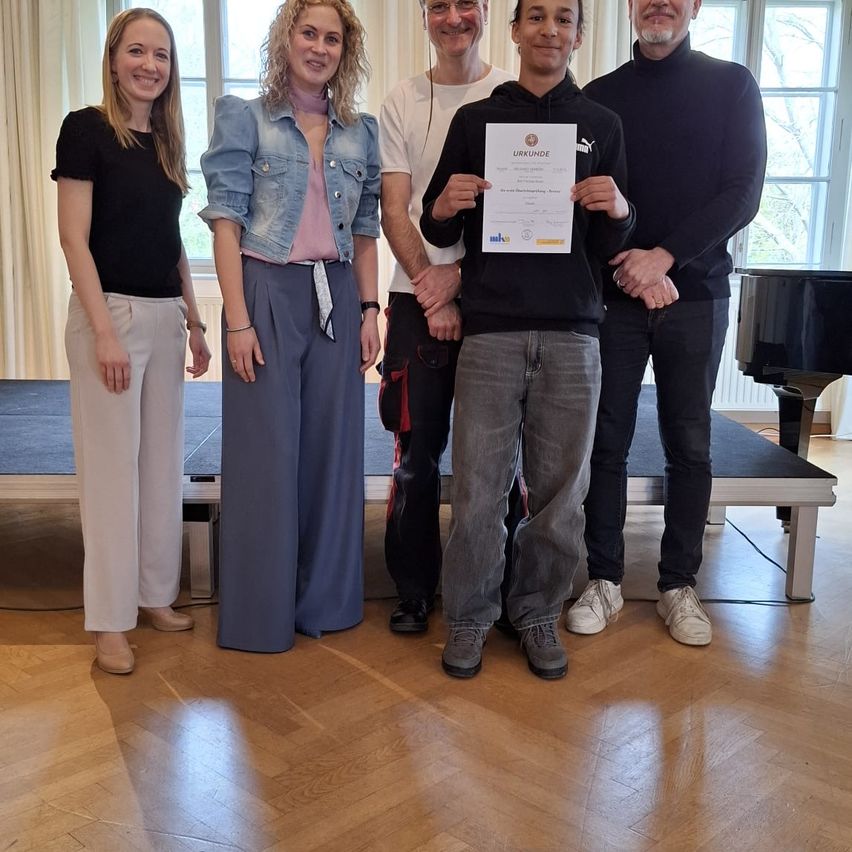 Fünf Personen stehen in einem Raum mit Holzboden zusammen. Ein Junge hält ein Zertifikat in beiden Händen. Hinter ihm stehen zwei Männer und zwei Frauen. Dahinter steht ein Klavier. Es gibt ein Fenster mit weißen Vorhängen.