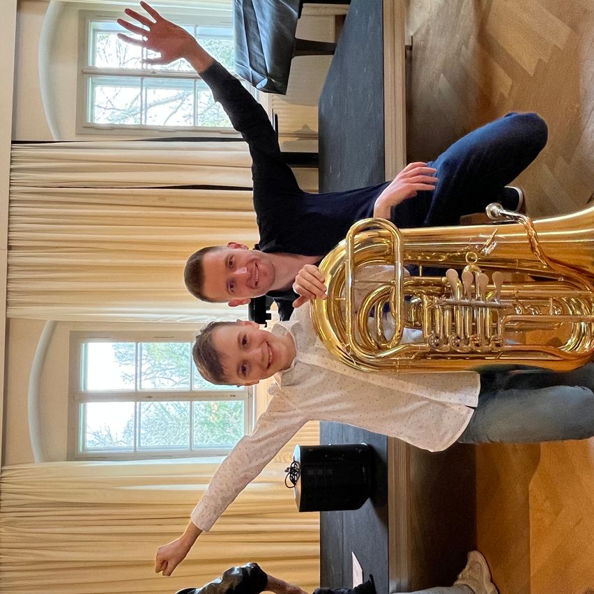 Ein Junge und ein Mann posieren mit einem Tuba in einem Raum mit großen Fenstern. Der Junge lächelt, während er die Tuba hält, und der Mann sitzt hinter ihm.