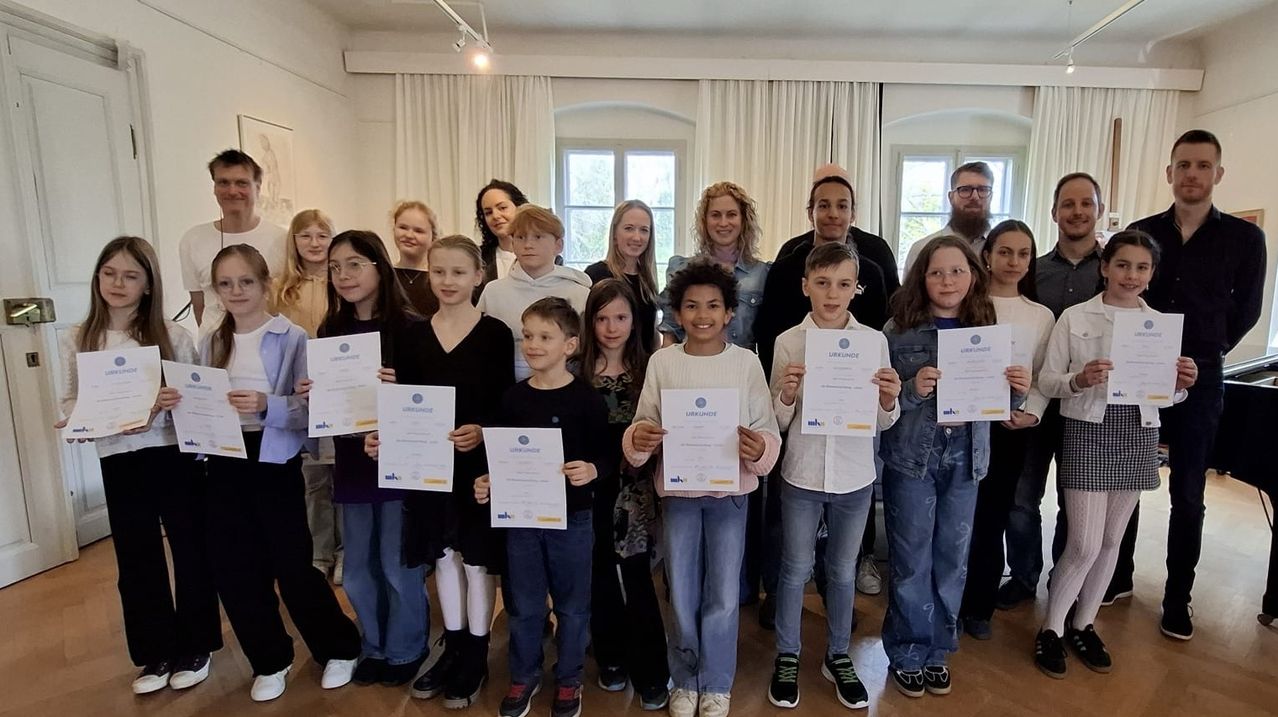 Eine Gruppe von Kindern und Erwachsenen steht zusammen und hält Urkunden in einem Raum mit Holzböden und großen Fenstern. Sie alle lächeln für die Kamera.