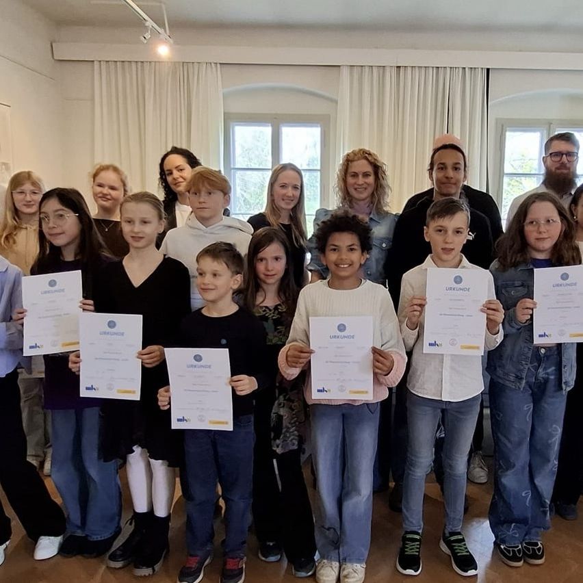 Eine Gruppe von Kindern und Erwachsenen steht zusammen und hält Urkunden in einem Raum mit Holzböden und großen Fenstern. Sie alle lächeln für die Kamera.