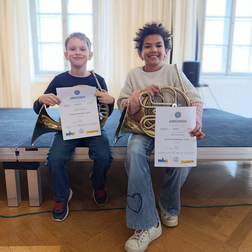 Zwei junge Jungen sitzen lächelnd auf einer Bühne und halten Urkunden in der Hand. Sie halten Blechblasinstrumente, einer hält ein Waldhorn und der andere eine Trompete. Sie tragen beide Jeans und Sneakers. Hinter ihnen steht ein Lautsprecher, ein Heizkörper und ein blauer Teppich auf der Bühne.