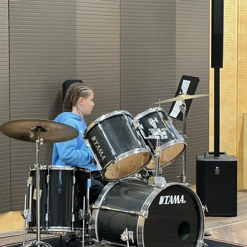Ein junger Mensch mit geflochtenen Haaren sitzt hinter einem Schlagzeug in einem Musikraum. Das Schlagzeug hat vier Trommeln und zwei Becken. Die Wände sind akustische Paneele.