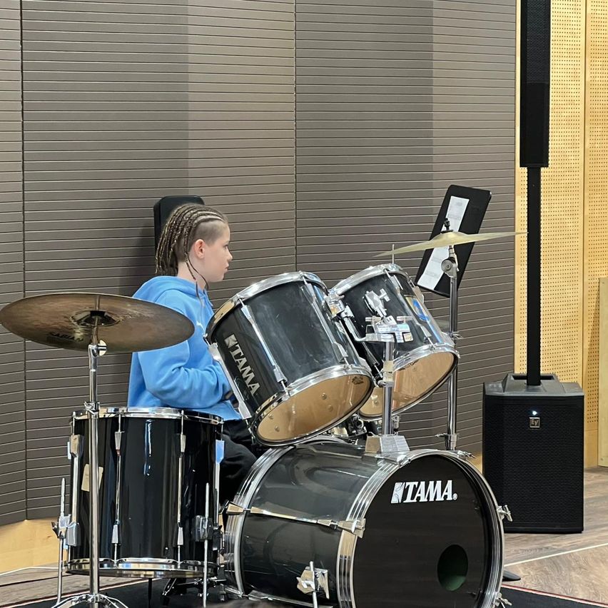 Ein junger Mensch mit geflochtenen Haaren sitzt hinter einem Schlagzeug in einem Musikraum. Das Schlagzeug hat vier Trommeln und zwei Becken. Die Wände sind akustische Paneele.