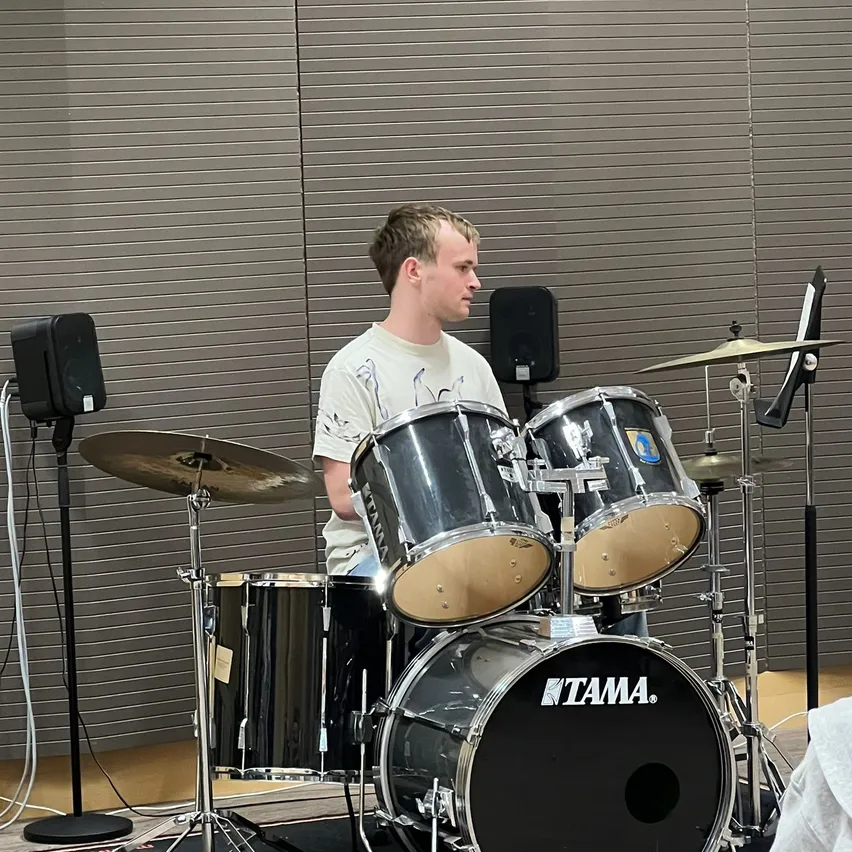 Ein Mann in einem weißen T-Shirt spielt eine Trommel mit einem Tama-Bass, Becken und Lautsprechern in einem Raum mit grauen Jalousien.