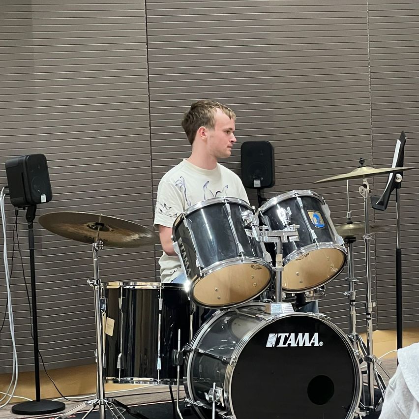 Ein Mann in einem weißen T-Shirt spielt eine Trommel mit einem Tama-Bass, Becken und Lautsprechern in einem Raum mit grauen Jalousien.