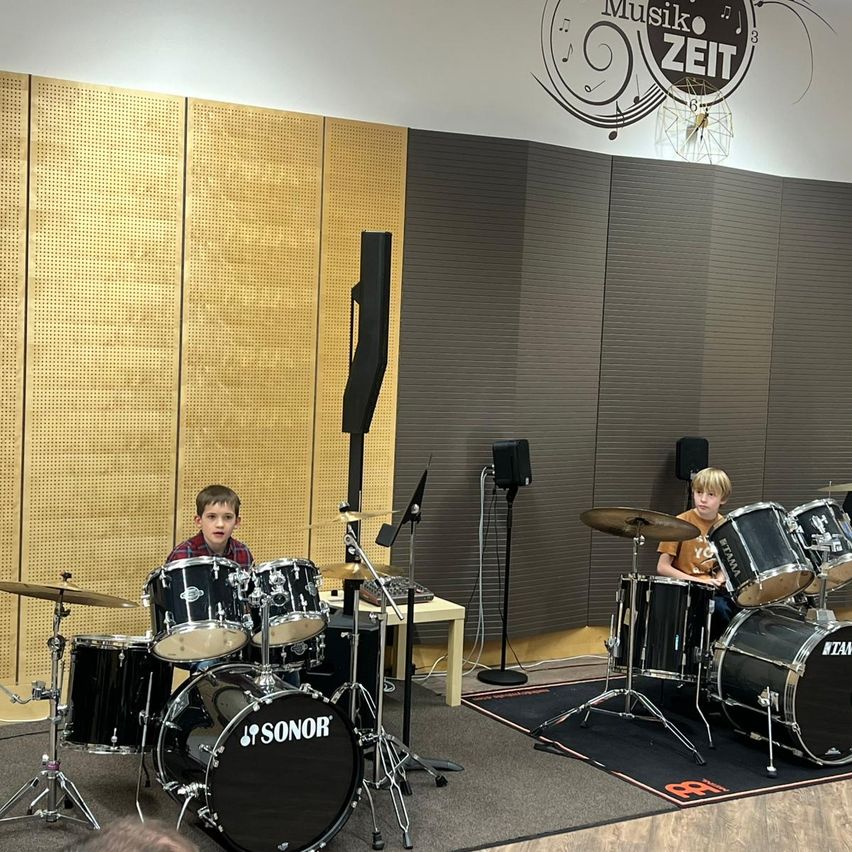 Zwei junge Jungen spielen Schlagzeug in einem Raum mit einer Wand, die das Wort 'Zeit' zeigt. Jeder Junge hat ein Schlagzeug vor sich mit Becken. Der Raum hat schallabsorbierende Paneele an der Wand.