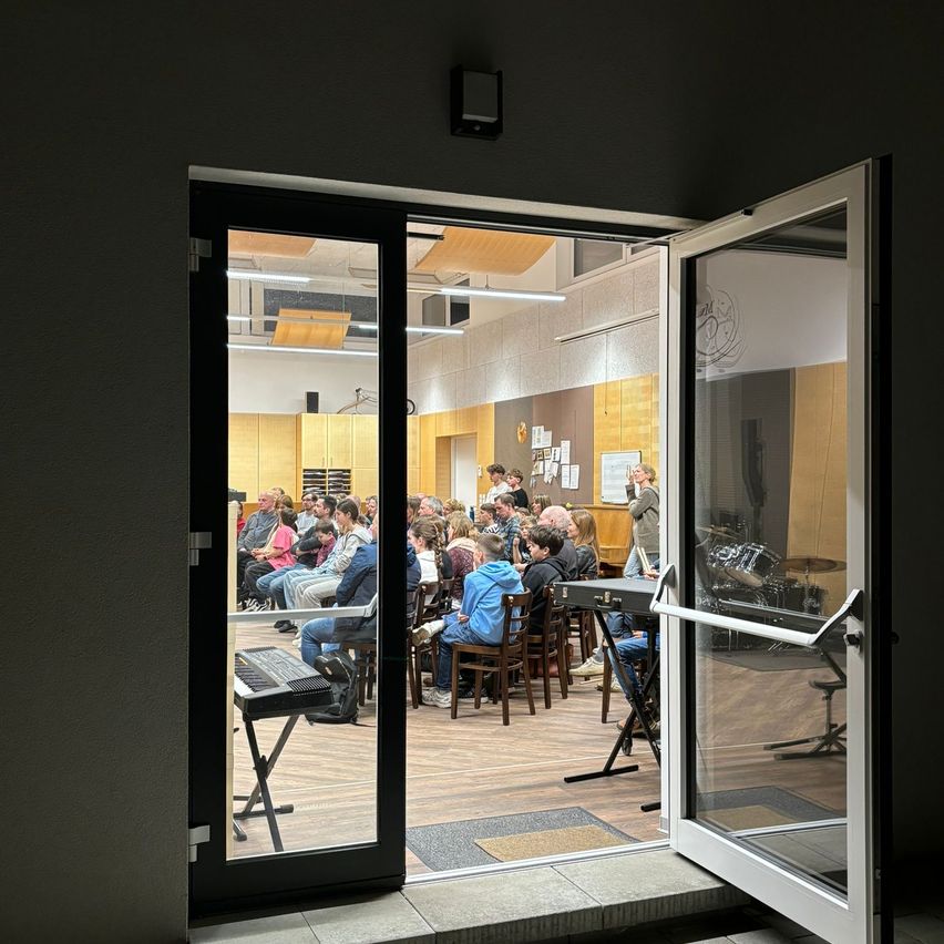 Eine Gruppe von Menschen sitzt in einem Raum mit Holzböden und Stühlen und hört einer Präsentation zu. Musikinstrumente sind sichtbar.