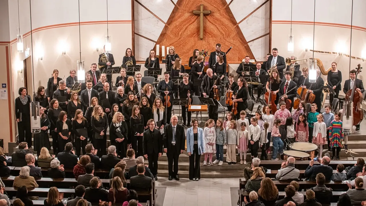 Ein großes Orchester steht in einer Kirche, mit Musikern, die verschiedene Instrumente spielen. Mehrere Kinder stehen vor ihnen, möglicherweise singend. Das Publikum sitzt im Vordergrund.