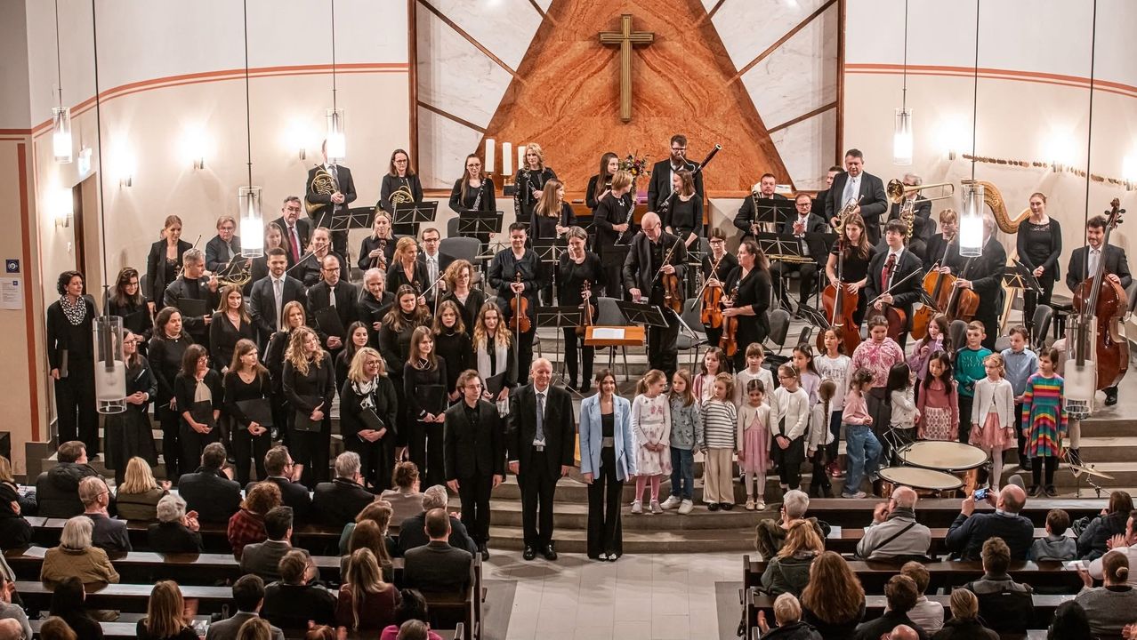 Ein großes Orchester steht in einer Kirche, mit Musikern, die verschiedene Instrumente spielen. Mehrere Kinder stehen vor ihnen, möglicherweise singend. Das Publikum sitzt im Vordergrund.