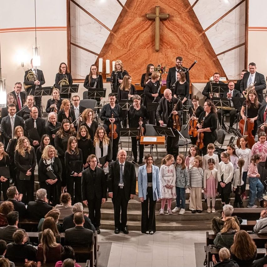 Ein großes Orchester steht in einer Kirche, mit Musikern, die verschiedene Instrumente spielen. Mehrere Kinder stehen vor ihnen, möglicherweise singend. Das Publikum sitzt im Vordergrund.