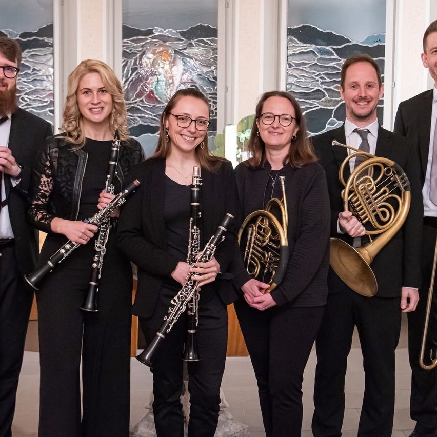 Fünf Personen in formeller Kleidung posieren für ein Foto mit ihren Musikinstrumenten. Zwei Frauen halten Klarinetten, drei Männer halten französische Hörner. Im Hintergrund sind Buntglasfenster zu sehen.