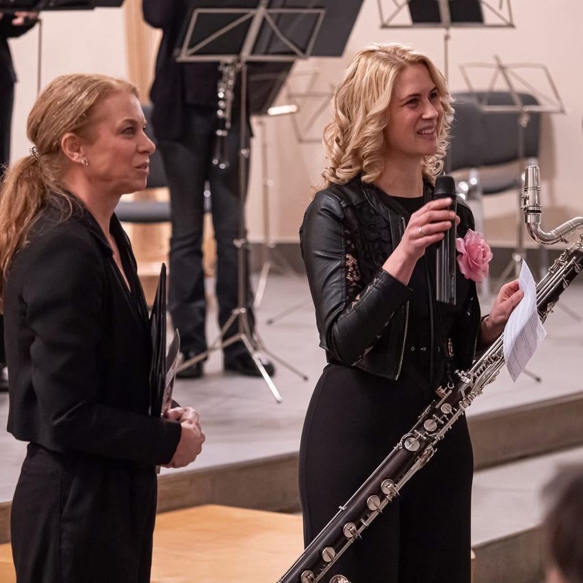 Eine Frau hält ein Mikrofon und ein Papier und lächelt. Daneben steht eine andere Frau mit einem Buch. Dahinter steht eine Person in der Nähe eines Notenständers.