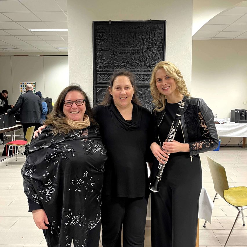 Drei Frauen in schwarzen Kleidern stehen lächelnd in einem Raum. Eine Frau hält eine Klarinette. Hinter ihnen steht eine schwarze Skulptur, und im Hintergrund sind Menschen zu sehen.