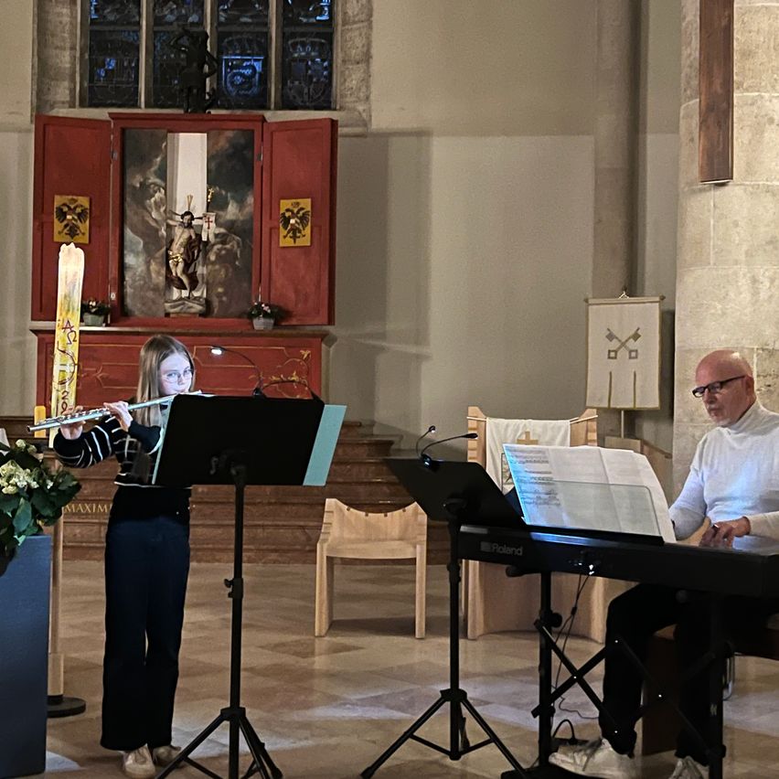 Ein junges Mädchen spielt die Flöte, während ein Mann sie am Keyboard begleitet. Sie befinden sich beide in einer Kirche mit Musikinstrumenten und einem Blumenarrangement.