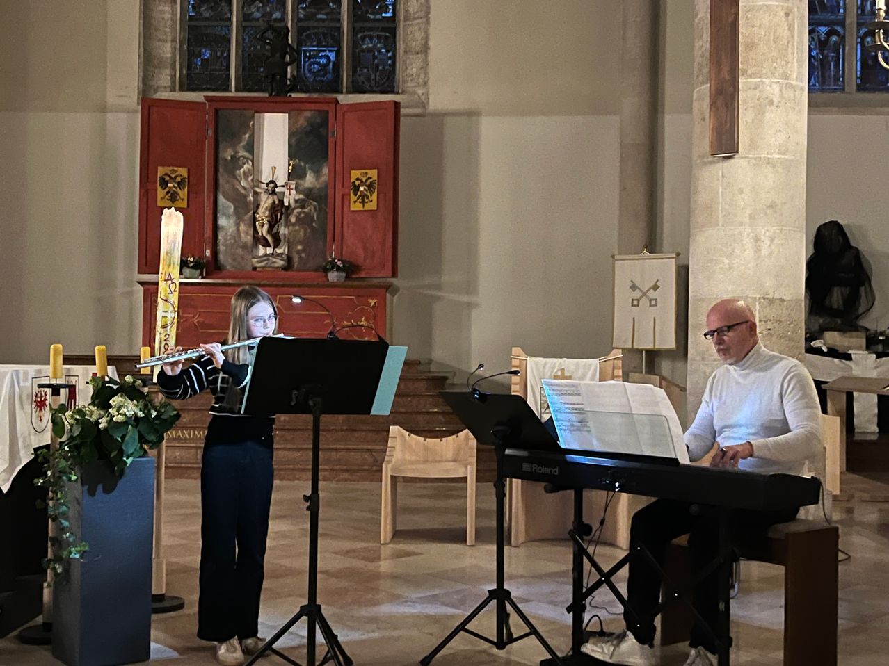 Ein junges Mädchen spielt die Flöte, während ein Mann sie am Keyboard begleitet. Sie befinden sich beide in einer Kirche mit Musikinstrumenten und einem Blumenarrangement.