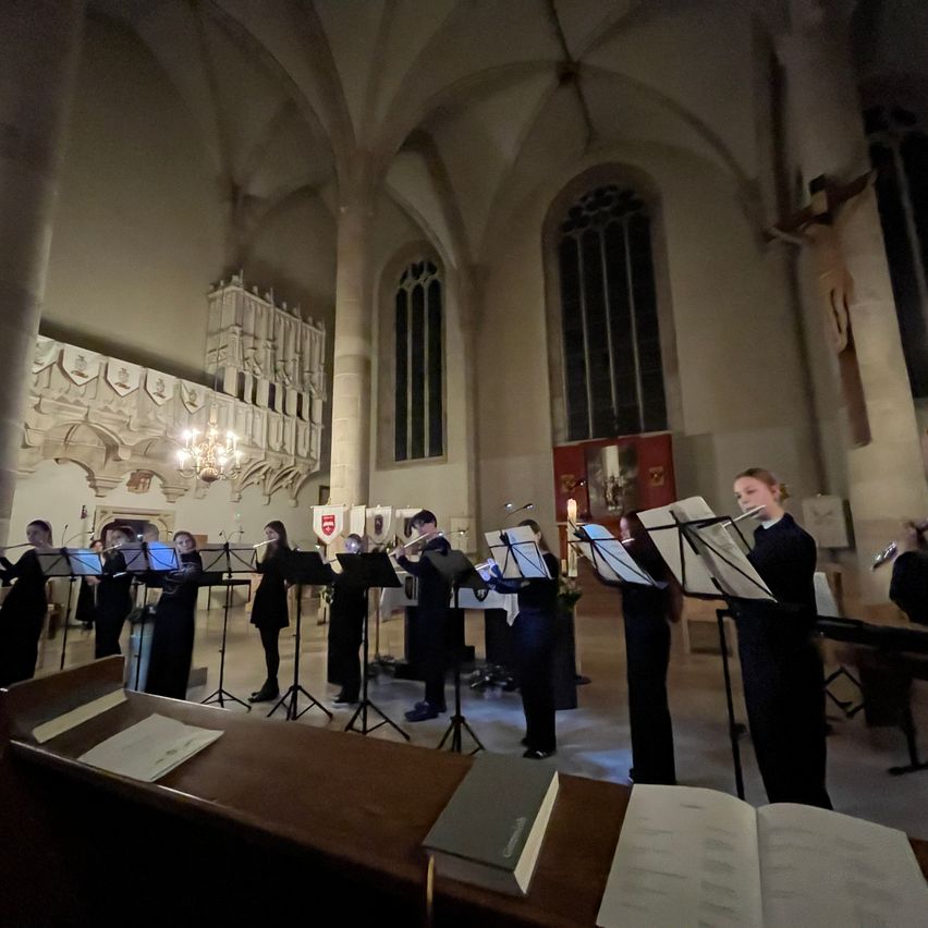 Eine Gruppe von Musikern in einer Kirchenumgebung, die Instrumente spielen. Sie stehen vor Notenständern mit offenen Büchern.