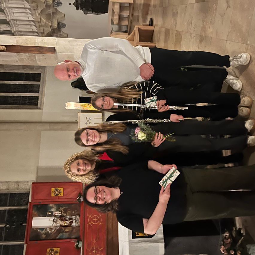 Eine Gruppe von vier Personen, zwei Männer und zwei Frauen, posiert für ein Foto in einer Kirche. Die Frauen halten Blumen und Flöten, während ein Mann ein Geschenk hält. Sie stehen in der Nähe einer roten Wand mit einem Gemälde.