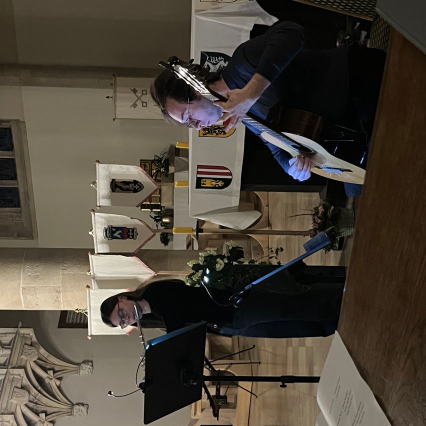 Zwei Personen spielen Gitarren in einer Kirchenumgebung. Eine Person steht auf der Bühne, die andere darunter, beide umgeben von Blumen und dekorativen Bannern.