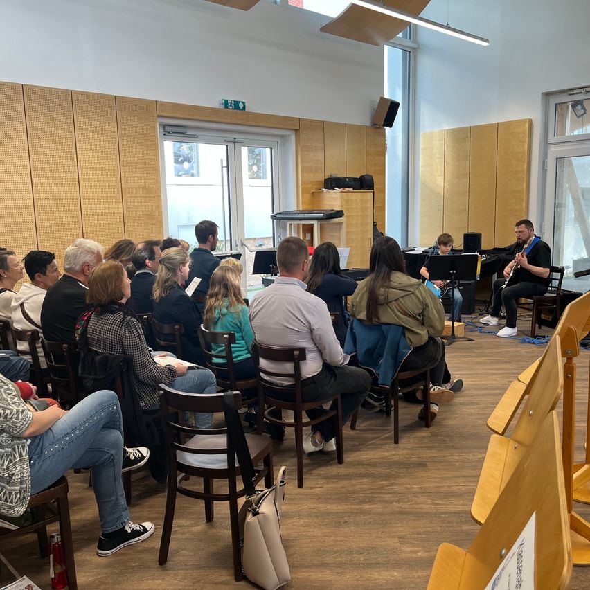 Eine Gruppe von Menschen sitzt in einem Raum und hört einem Mann zu, der Gitarre spielt. Einige Musikinstrumente stehen vor ihnen.