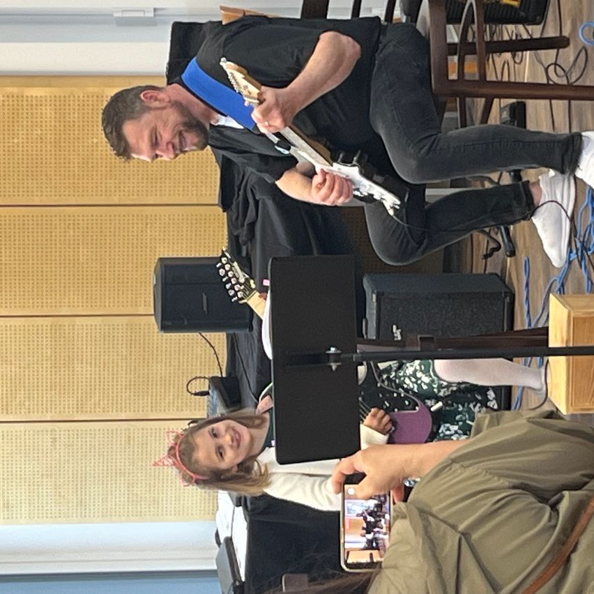 Ein Mann spielt eine E-Gitarre, während ein Mädchen und eine Frau zusehen und ein Handy aufnimmt.