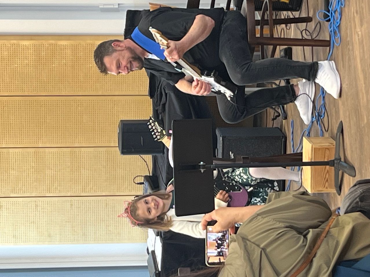 Ein Mann spielt eine E-Gitarre, während ein Mädchen und eine Frau zusehen und ein Handy aufnimmt.