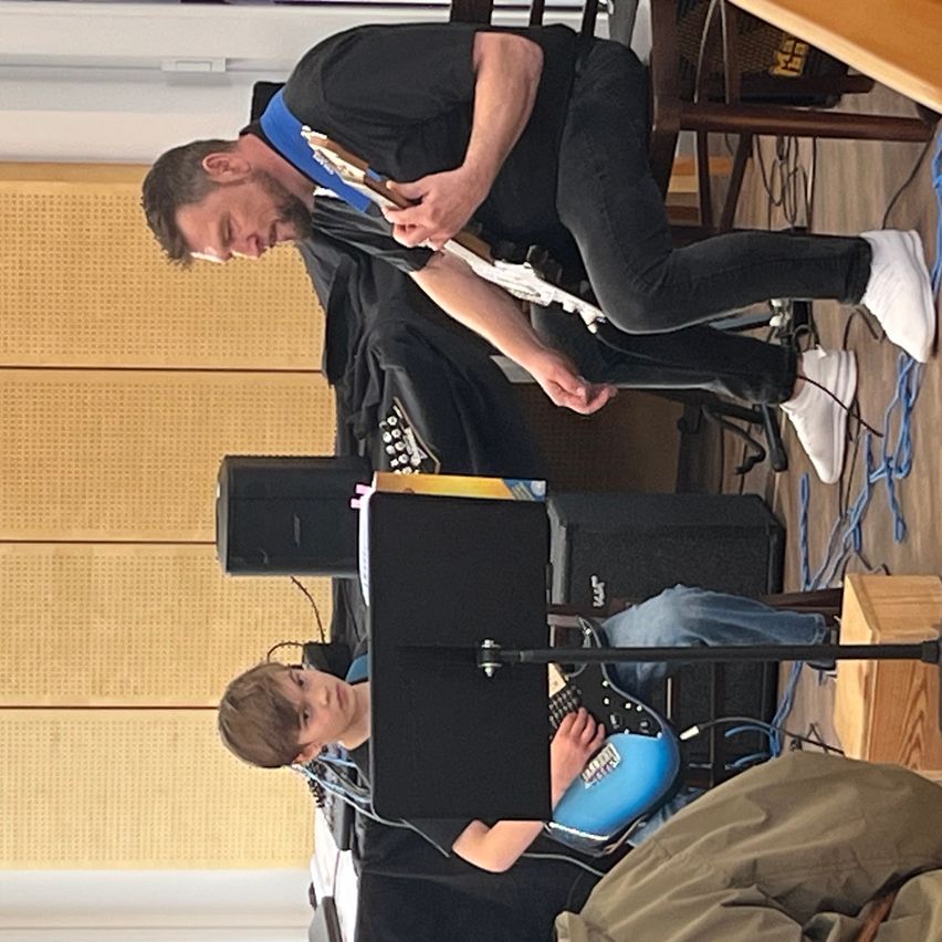 Zwei Männer spielen Gitarre. Einer steht, der andere sitzt. Sie sind in einem Raum mit einer Holzwand und einem schwarzen Lautsprecher.