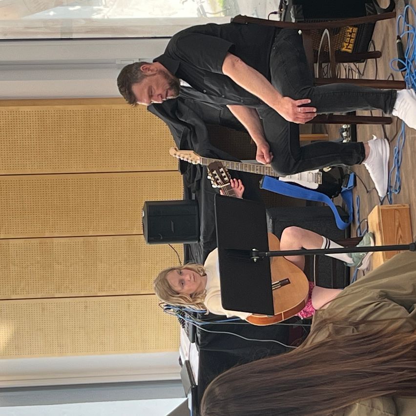 Ein Mann und ein Mädchen spielen Gitarren in einem Raum mit Holzboden und Fenster. Der Mann sitzt auf einem Stuhl, während das Mädchen auf dem Boden sitzt.