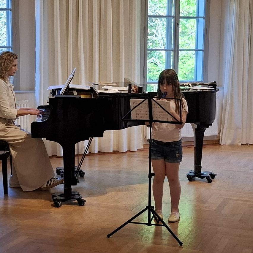 Eine Frau spielt Klavier, während ein junges Mädchen in ein Mikrofon singt, vor einem Notenständer in einem Raum mit großen Fenstern und Holzböden.