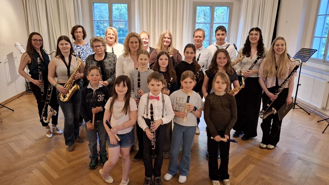 Eine Gruppe von Kindern und Erwachsenen steht lächelnd in einem Raum zusammen. Die Kinder halten Musikinstrumente wie Flöten, Klarinetten und Saxophone. Die Erwachsenen, darunter Frauen und Männer, stehen hinter den Kindern und lächeln ebenfalls, während sie ebenfalls Instrumente halten.