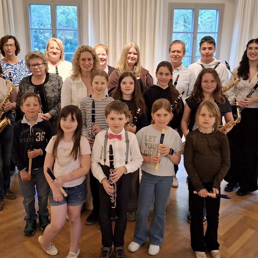 Eine Gruppe von Kindern und Erwachsenen steht lächelnd in einem Raum zusammen. Die Kinder halten Musikinstrumente wie Flöten, Klarinetten und Saxophone. Die Erwachsenen, darunter Frauen und Männer, stehen hinter den Kindern und lächeln ebenfalls, während sie ebenfalls Instrumente halten.