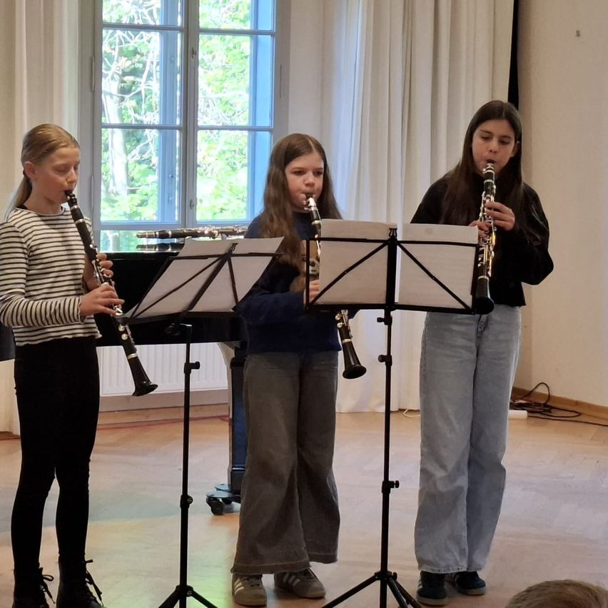 Drei junge Mädchen spielen Klarinetten in einem Raum mit Klavier und weißen Vorhängen. Ein Mädchen trägt ein gestreiftes Shirt, während die anderen lässige Kleidung tragen.