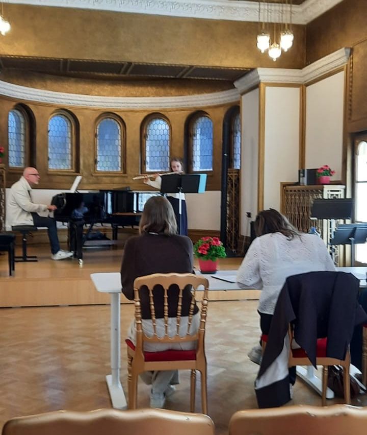 Eine Frau spielt Flöte auf einer Bühne mit einem Klavier dahinter. Zwei Personen sitzen vor einem Schreibtisch und lauschen aufmerksam. Der Raum hat Bogenfenster und eine Kronleuchter.