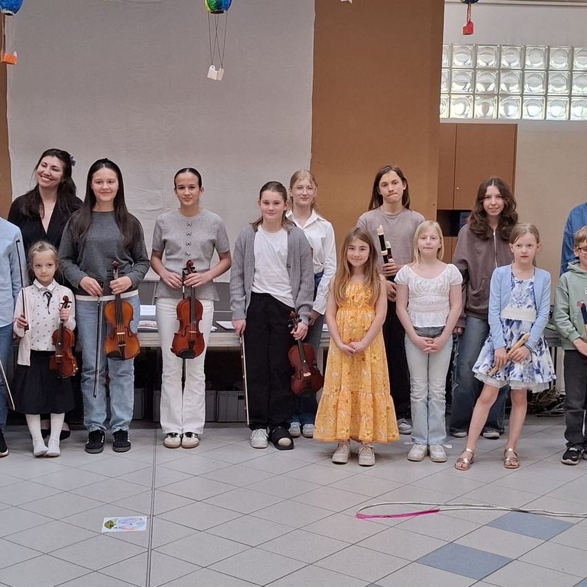 Eine Gruppe junger Musiker, sowohl Jungen als auch Mädchen, posiert für ein Foto mit ihren Instrumenten. Sie stehen in einer Reihe, einige halten Violinen und andere Bögen. Sie scheinen bei einer Musikveranstaltung zu sein.