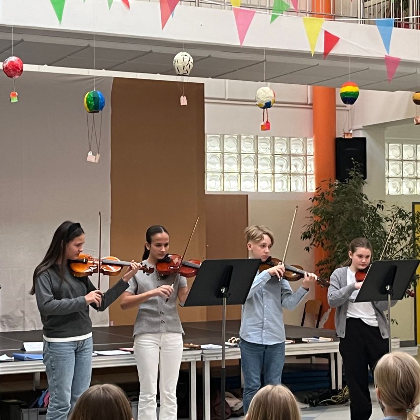 Vier junge Geiger spielen auf der Bühne vor einem Publikum. Die Bühne ist mit Wimpelgirlanden und Luftballons geschmückt. Dahinter befindet sich eine Wand mit Glaselementen.