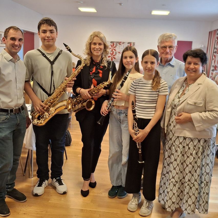 Eine Gruppe von Menschen steht lächelnd für ein Foto in einem Raum. Zwei von ihnen halten Musikinstrumente, eines ein Saxophon und das andere eine Klarinette.
