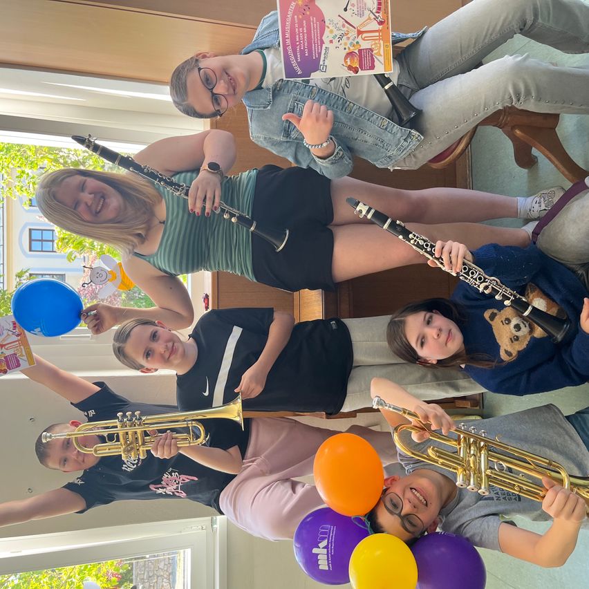 Gruppe von Kindern, die lächeln und Musikinstrumente halten. Luftballons im Hintergrund. Ein Kind hält eine Trompete, ein anderes eine Klarinette. Ein Mädchen liest ein Buch.