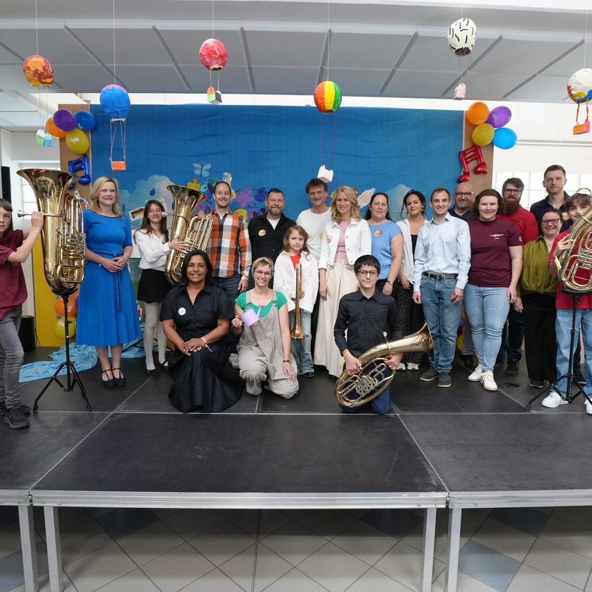 Eine Gruppe von Menschen posiert mit Blasinstrumenten auf einer Bühne, mit einem farbenfrohen Hintergrund und Luftballons.