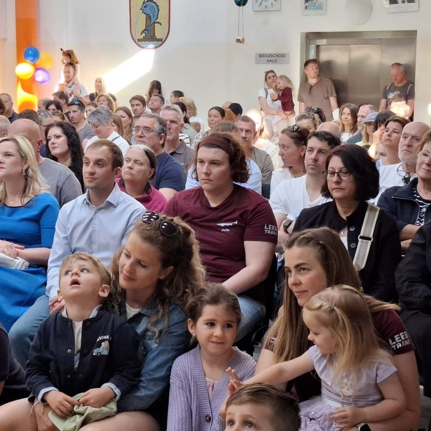 Eine große Menschenmenge versammelt sich in einem Saal, einige stehen und andere sitzen. Der Raum ist mit Luftballons gefüllt, und es gibt ein Schild an der Wand. Die Menschenmenge ist in Alter und Geschlecht vielfältig.