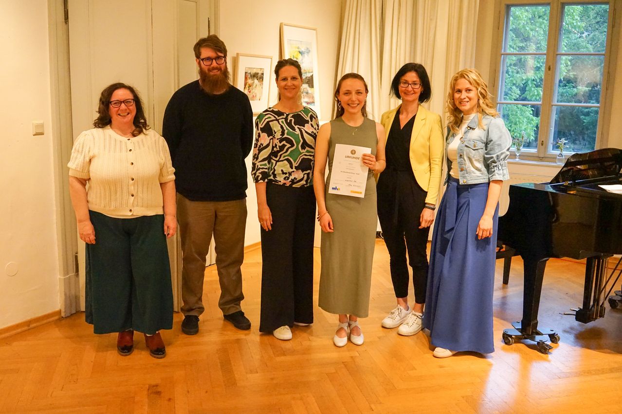 Eine Gruppe von sechs Personen, darunter eine Frau mit einem Zertifikat, posiert für ein Foto in einem Raum. Sie alle lächeln.